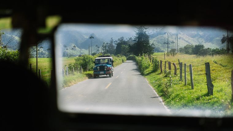 A rear-view mirror showing a jeep on a quiet country road
